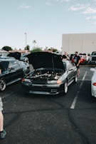 A car meet-up in a parking lot features several vehicles with their hoods open. In the center, a dark-colored car displays its engine for viewing. People are gathered around the cars, some engaged in conversation. The setting includes a clear sky and a few palm trees in the background.