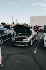 A car meet-up in a parking lot features several vehicles with their hoods open. In the center, a dark-colored car displays its engine for viewing. People are gathered around the cars, some engaged in conversation. The setting includes a clear sky and a few palm trees in the background.