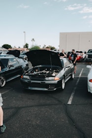 A car meet-up in a parking lot features several vehicles with their hoods open. In the center, a dark-colored car displays its engine for viewing. People are gathered around the cars, some engaged in conversation. The setting includes a clear sky and a few palm trees in the background.