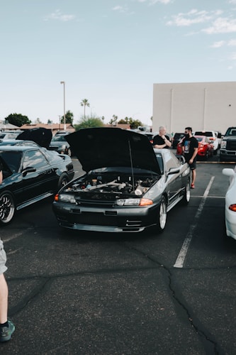 A car meet-up in a parking lot features several vehicles with their hoods open. In the center, a dark-colored car displays its engine for viewing. People are gathered around the cars, some engaged in conversation. The setting includes a clear sky and a few palm trees in the background.