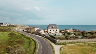 A coastal scene featuring a large, elegant house with a dark roof, situated beside a curving road that runs parallel to the ocean. The property is surrounded by lush greenery and well-maintained garden areas. Behind the house, the expansive ocean is visible, with clear skies overhead. Several smaller buildings and houses are situated further along the coastline.