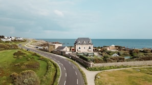A coastal scene featuring a large, elegant house with a dark roof, situated beside a curving road that runs parallel to the ocean. The property is surrounded by lush greenery and well-maintained garden areas. Behind the house, the expansive ocean is visible, with clear skies overhead. Several smaller buildings and houses are situated further along the coastline.
