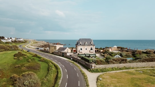 A coastal scene featuring a large, elegant house with a dark roof, situated beside a curving road that runs parallel to the ocean. The property is surrounded by lush greenery and well-maintained garden areas. Behind the house, the expansive ocean is visible, with clear skies overhead. Several smaller buildings and houses are situated further along the coastline.