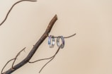A pair of colorful gemstone rings rests on a dark, twisted tree branch against a neutral beige background. The rings have various stones in shades of blue, pink, and purple, creating a vibrant contrast with the natural texture of the branch.