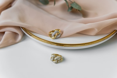 A close-up of elegant jewelry including a gold and silver ring resting on a white plate with a gold rim. Soft beige fabric drapes over the plate, adding a touch of luxury. A small green branch accompanies the setup, enhancing the sophisticated look.