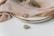 A close-up of elegant jewelry including a gold and silver ring resting on a white plate with a gold rim. Soft beige fabric drapes over the plate, adding a touch of luxury. A small green branch accompanies the setup, enhancing the sophisticated look.