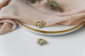 A close-up of elegant jewelry including a gold and silver ring resting on a white plate with a gold rim. Soft beige fabric drapes over the plate, adding a touch of luxury. A small green branch accompanies the setup, enhancing the sophisticated look.