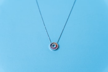 A delicate necklace with a circular pendant adorned with gemstones in a range of colors including red, yellow, and blue, set against a light blue background. The chain is thin and appears to be made of a silver-colored metal.