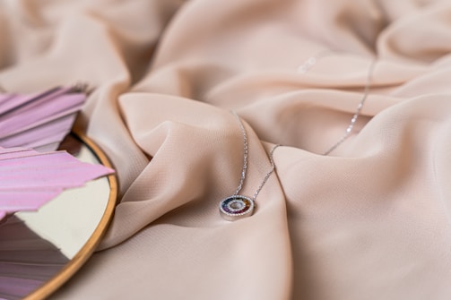 A delicate pendant necklace with a circular design lies on a soft beige fabric. The pendant features a colorful arrangement of small gemstones. In the left corner, part of a circular handheld fan with soft pink pleats is visible, reflecting slightly on a mirrored surface.
