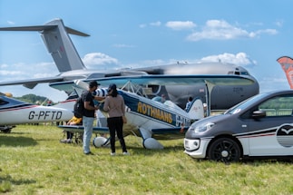 An outdoor scene features several aircrafts on display, including a large military cargo plane in the background and a smaller vintage biplane in the foreground. A car is parked to the right. Two people stand near the biplane, engaged in conversation, with one pointing towards it. The setting is grassy with a clear blue sky overhead, suggesting a daytime event, potentially an airshow.