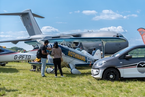 An outdoor scene features several aircrafts on display, including a large military cargo plane in the background and a smaller vintage biplane in the foreground. A car is parked to the right. Two people stand near the biplane, engaged in conversation, with one pointing towards it. The setting is grassy with a clear blue sky overhead, suggesting a daytime event, potentially an airshow.