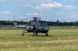 A military helicopter marked with 'ROYAL NAVY' is parked on a grassy airfield. The helicopter has the number 426 printed on it and features a distinct dark color with a rotor on top. The background includes a clear sky with scattered clouds and a tree line separating the field from the horizon.