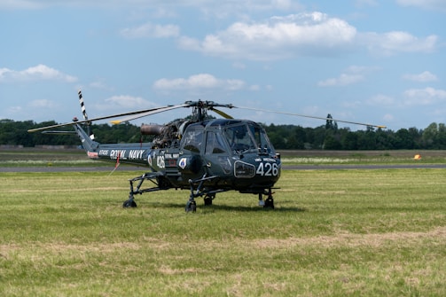A military helicopter marked with 'ROYAL NAVY' is parked on a grassy airfield. The helicopter has the number 426 printed on it and features a distinct dark color with a rotor on top. The background includes a clear sky with scattered clouds and a tree line separating the field from the horizon.