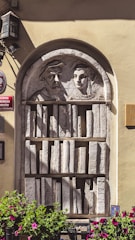 A stone relief sculpture is embedded in a wall, featuring two human figures wearing what appear to be traditional Renaissance clothing. Below the figures is a carved bookshelf filled with intricately detailed book spines. The scene is framed by an arch and flanked by potted plants with vibrant purple flowers.