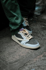 A person is wearing stylish sneakers with beige laces and a combination of dark and light blue colors. The shoes are on a concrete surface, and the person is wearing green pants.