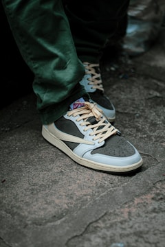 A person is wearing stylish sneakers with beige laces and a combination of dark and light blue colors. The shoes are on a concrete surface, and the person is wearing green pants.