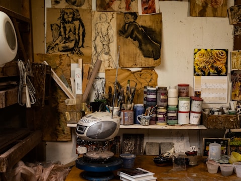 An artist's workspace filled with various materials, including paint jars, brushes, and art supplies. There are paintings and sketches on the walls, some depicting human figures. A radio sits on the counter amidst the cluttered yet creative environment.