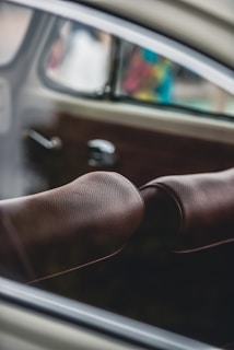 A close-up view of the interior of a car, featuring brown perforated leather upholstery on the seat. The window provides a blurred view of the outside with hints of color, suggesting a busy or colorful environment outside.