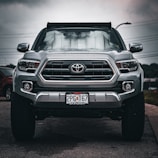 A front-facing silver Toyota pickup truck is parked on the street with a sunshade on the windshield and a visible license plate. The background includes a cloudy sky and other parked cars.