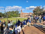 A crowd of people gathers around a sports field on a sunny day, with some wearing casual clothes and holding snacks. In the background, a historic brick building with a clock tower and lush greenery can be seen.
