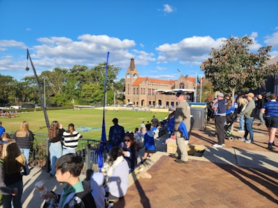A crowd of people gathers around a sports field on a sunny day, with some wearing casual clothes and holding snacks. In the background, a historic brick building with a clock tower and lush greenery can be seen.