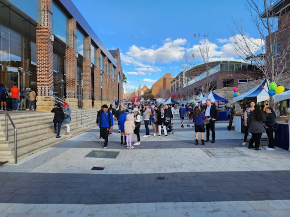 A bustling outdoor market scene with numerous people walking along a paved walkway framed by vendor stalls with blue and white canopies. The market is set against a backdrop of modern brick buildings with large glass windows. Balloons add a festive touch to the stalls, and leafless trees line the path under a vibrant blue sky with scattered clouds.