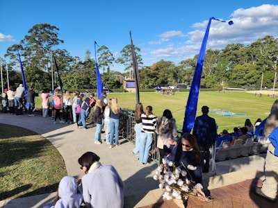 A group of people are gathered around the edge of a sports field, some standing and others seated. The field is surrounded by tall trees and a clear blue sky. Some individuals appear to be spectating, while others are participating in an event on the field. Blue flags are visible and fluttering in the breeze.