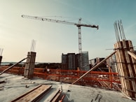 A construction site featuring a tall crane in the foreground with various building materials scattered around. Unfinished concrete pillars with exposed rebar are visible, and several tall buildings are in the background under a clear sky.