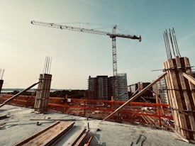 A construction site featuring a tall crane in the foreground with various building materials scattered around. Unfinished concrete pillars with exposed rebar are visible, and several tall buildings are in the background under a clear sky.