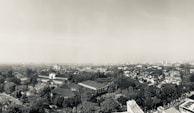 Aerial view of an urban landscape with a blend of modern and colonial-style buildings surrounded by numerous trees and greenery. The city unfolds into the distance, with a hazy sky above and scattered high-rise buildings visible in the background. The architecture and layout suggest a harmonious integration of urban development and nature.