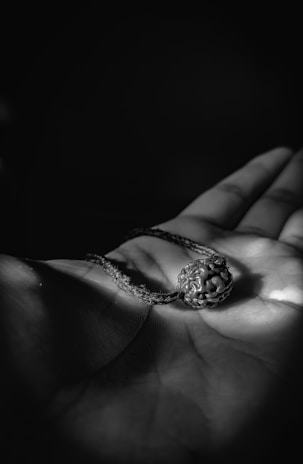 A hand is holding a rudraksha bead with a string attached. The image is in black and white, highlighting the intricate textures of the bead and the creases on the palm.