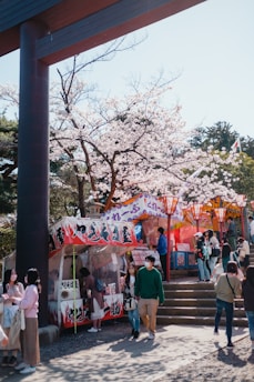 A joyful travel vlogger interacting with locals while exploring a bustling street market under soft pink sakura petals.
