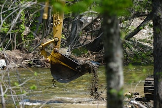 A construction excavator arm with a yellow hydraulic excavator bucket dips into a clear, shallow river surrounded by dense forest. The surrounding environment is lush with green leaves and trees, creating a natural setting.