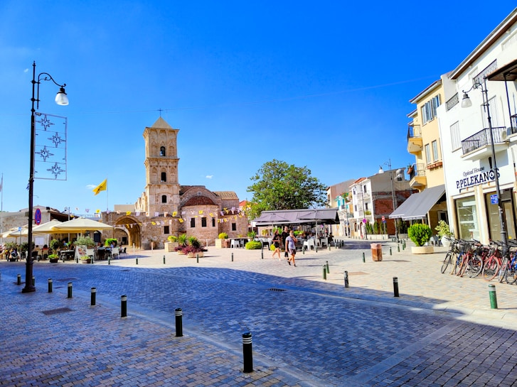 A sunny plaza with a historic stone church building featuring a bell tower surrounded by flags and banners. The scene includes people walking, outdoor café seating with umbrellas, bicycles parked on the side, and a mix of stone and modern buildings.