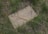 A gravestone marked with the word 'UNKNOWN' lies among tall grass and scattered small purple flowers. Two dried roses rest diagonally across the stone, suggesting a sense of remembrance.
