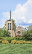 A stone church with a tall spire and a large, circular stained glass window is surrounded by lush greenery. The building's architecture features classic Gothic elements, and the well-maintained lawn and bushes add to the serene ambiance. The sky above is partly cloudy, complementing the peaceful scene.