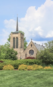A stone church with a tall spire and a large, circular stained glass window is surrounded by lush greenery. The building's architecture features classic Gothic elements, and the well-maintained lawn and bushes add to the serene ambiance. The sky above is partly cloudy, complementing the peaceful scene.