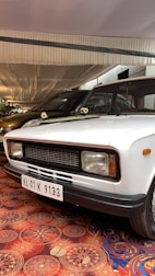 A white vintage car is parked indoors under a fabric-covered ceiling. The car has a black grille and rectangular headlights. The license plate displays the code KL 01 K 9133. Floral patterned carpet covers the floor, adding a decorative touch to the setting.