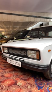 A white vintage car is parked indoors under a fabric-covered ceiling. The car has a black grille and rectangular headlights. The license plate displays the code KL 01 K 9133. Floral patterned carpet covers the floor, adding a decorative touch to the setting.