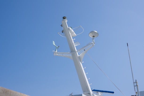 A white ship's mast extends vertically against a clear blue sky. The mast is equipped with various navigational and communication instruments, including antennas, a radar dome, and a small anemometer. Metallic ladders and railings are attached to the structure, indicating accessibility for maintenance.