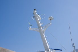 A white ship's mast extends vertically against a clear blue sky. The mast is equipped with various navigational and communication instruments, including antennas, a radar dome, and a small anemometer. Metallic ladders and railings are attached to the structure, indicating accessibility for maintenance.