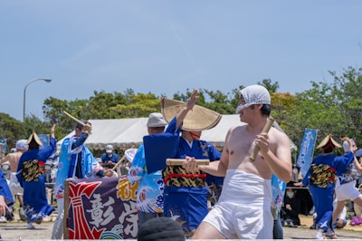 A group of people dressed in traditional Japanese festival attire, with some wearing blue kimonos and straw hats, are participating in a cultural celebration. One individual in the foreground is wearing minimal clothing and holding a wooden stick. Banners with Japanese characters and a tent are visible in the background, surrounded by green trees under a clear blue sky.
