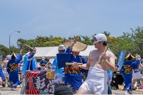 A group of people dressed in traditional Japanese festival attire, with some wearing blue kimonos and straw hats, are participating in a cultural celebration. One individual in the foreground is wearing minimal clothing and holding a wooden stick. Banners with Japanese characters and a tent are visible in the background, surrounded by green trees under a clear blue sky.