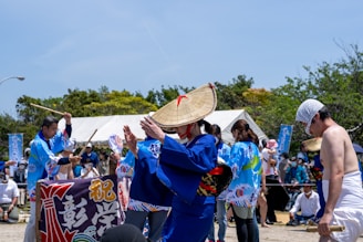 A cultural festival scene with people wearing traditional Japanese attire. A central figure is dressed in a blue kimono and a conical straw hat, engaging in a dance or performance. Others in similar blue attire hold wooden sticks and fans. A tent and banners are visible in the background, with spectators seated or standing around.