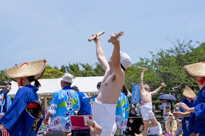 A group of people are wearing traditional clothing during what appears to be a cultural or festival celebration. Some are dressed in blue outfits with straw hats, while a man in the center raises wooden sticks with enthusiasm. The event takes place outdoors under a bright sky, and spectators can be seen in the background.