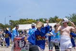 A group of people engaging in a festival or cultural event, wearing traditional clothing such as kimonos and straw hats. The scene includes dancers and musicians performing, with one man in minimal traditional attire holding sticks. The background shows tents and other observers, some of whom are clapping or taking photos.