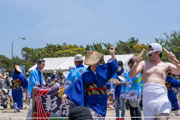 A group of people engaging in a festival or cultural event, wearing traditional clothing such as kimonos and straw hats. The scene includes dancers and musicians performing, with one man in minimal traditional attire holding sticks. The background shows tents and other observers, some of whom are clapping or taking photos.