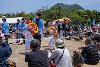 A lively outdoor festival setting with performers dressed in traditional Japanese attire. The performers, including children, are holding colorful red and yellow umbrellas and wearing face coverings. They are surrounded by an audience seated on the ground, many wearing hats and some holding cameras. Trees and a mountain are visible in the background under a clear blue sky.