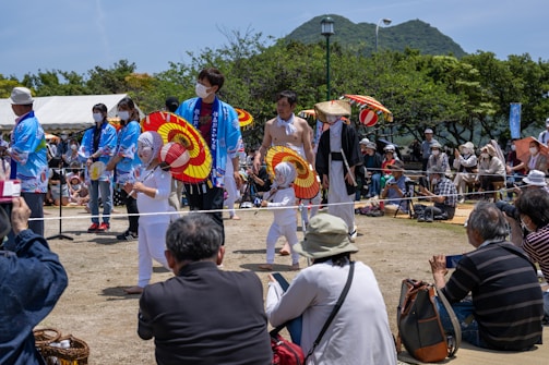 A lively outdoor festival setting with performers dressed in traditional Japanese attire. The performers, including children, are holding colorful red and yellow umbrellas and wearing face coverings. They are surrounded by an audience seated on the ground, many wearing hats and some holding cameras. Trees and a mountain are visible in the background under a clear blue sky.