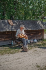 An elderly man sits on a stool playing a stringed musical instrument in a rustic outdoor setting. He wears a light-colored hat and shirt. Behind him, a wooden structure provides a backdrop with trees and greenery visible in the background. On the ground near him, there is a woven bag and some artwork displayed.